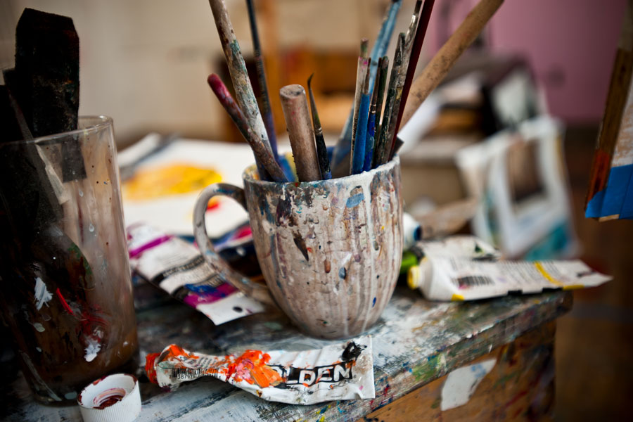 Messy art supplies on a table. Dirty paint brushes are resting in a white mug covered in paint.